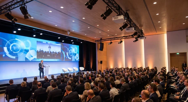 Salle de conférence ou d'assemblée générale professionnelle moderne, avec une grande audience assise face à une scène. Au centre : un orateur à un pupitre, écran LED géant haute définition en arrière-plan affichant des contenus (graphiques, vidéo, présentation). Projecteurs de scène professionnels, vidéoprojecteurs visibles en hauteur. Système de visioconférence intégré avec caméra motorisée pointée sur l'orateur. Public captif, configuration de type assemblée ou séminaire corporate. Photographie réaliste, éclairage professionnel chaud, plan large capturant la configuration technique et l'interaction humaine.